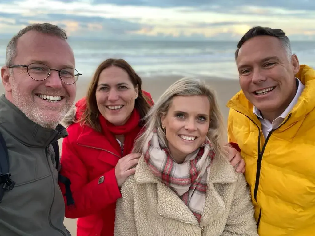 Vier mensen van Mens & Relatie lachen samen op het strand, met een grijze lucht en zee op de achtergrond.