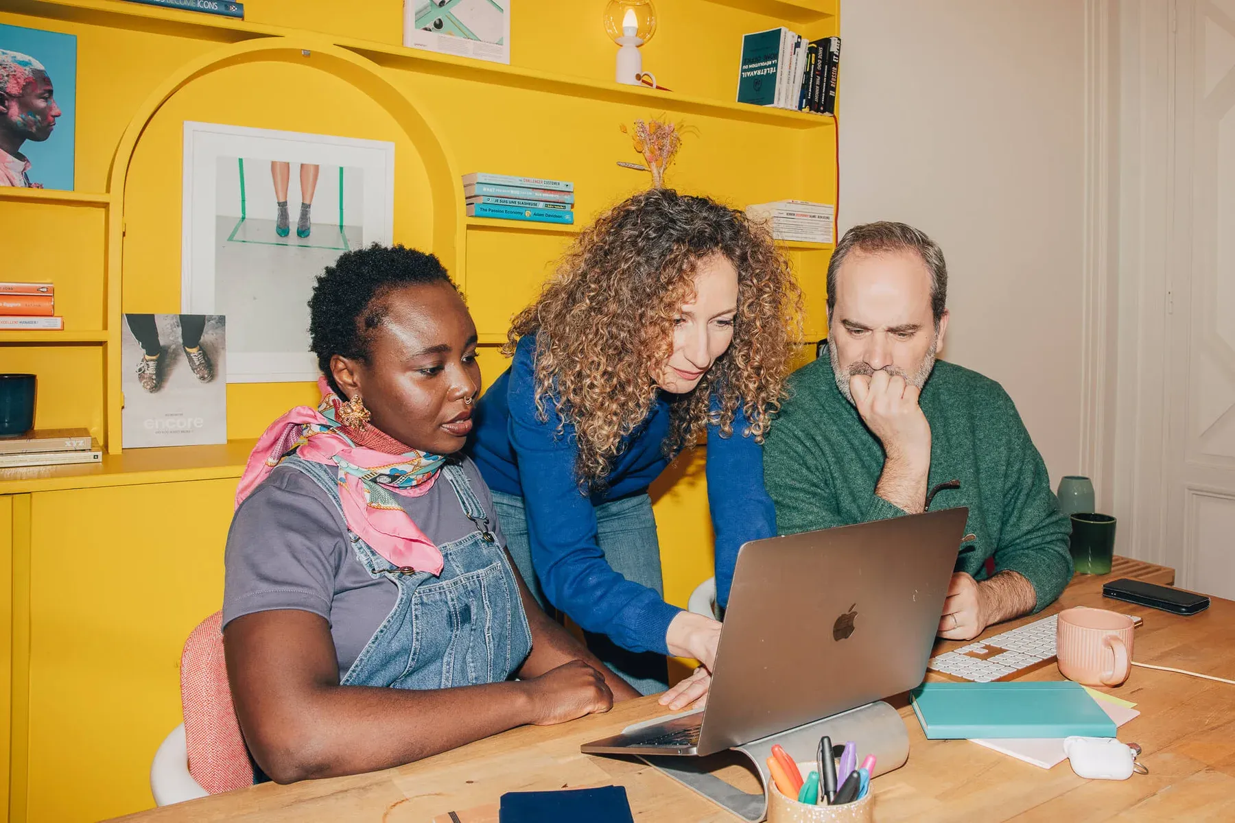 Trois collègues autour d'un ordinateur portable, dans un bureau aux étagères jaune vif